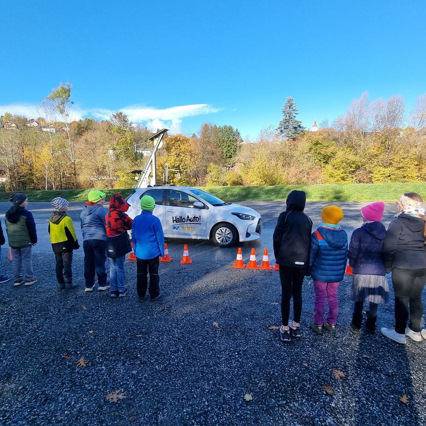 Eine Gruppe von Kindern steht um ein weißes Auto mit der Aufschrift 'Hallo Auto!' herum. Sie tragen Winterkleidung. Der Bereich ist von orangefarbenen Verkehrskegeln umgeben. Im Hintergrund befinden sich Bäume und ein Hügel.