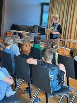 Eine Frau steht vor einer Gruppe von Kindern in einem Raum. Sie hält ein Buch in der Hand und liest laut vor. Die Kinder sitzen aufmerksam in Reihen von Stühlen. Der Raum hat einen Holzboden, ein Fenster und Vorhänge.