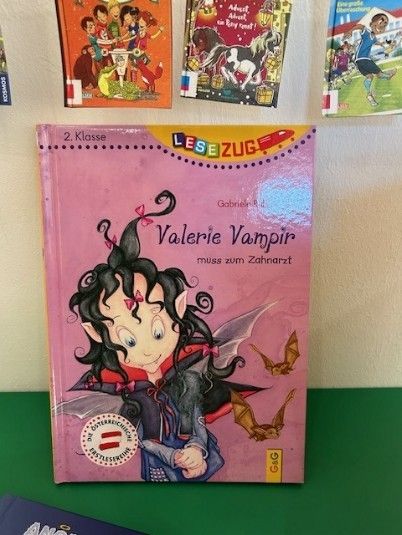 Ein rosafarbenes Buch mit dem Titel Valerie Vampir steht auf einer grünen Oberfläche mit anderen Büchern im Hintergrund. Das Cover zeigt ein Mädchen mit Fledermausflügeln und Vampirzähnen.