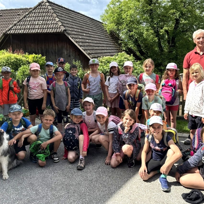 Eine Gruppe von Kindern, einige mit Mützen und Rucksäcken, posieren für ein Foto im Freien, mit einem Mann und einem Hund im Hintergrund.