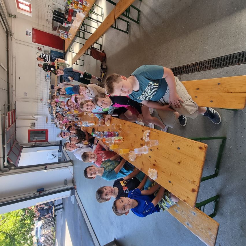 Eine Gruppe von Kindern sitzt mit Getränken um einen Holztisch, lächelt und befindet sich in einem Raum. Es gibt andere Personen im Hintergrund.