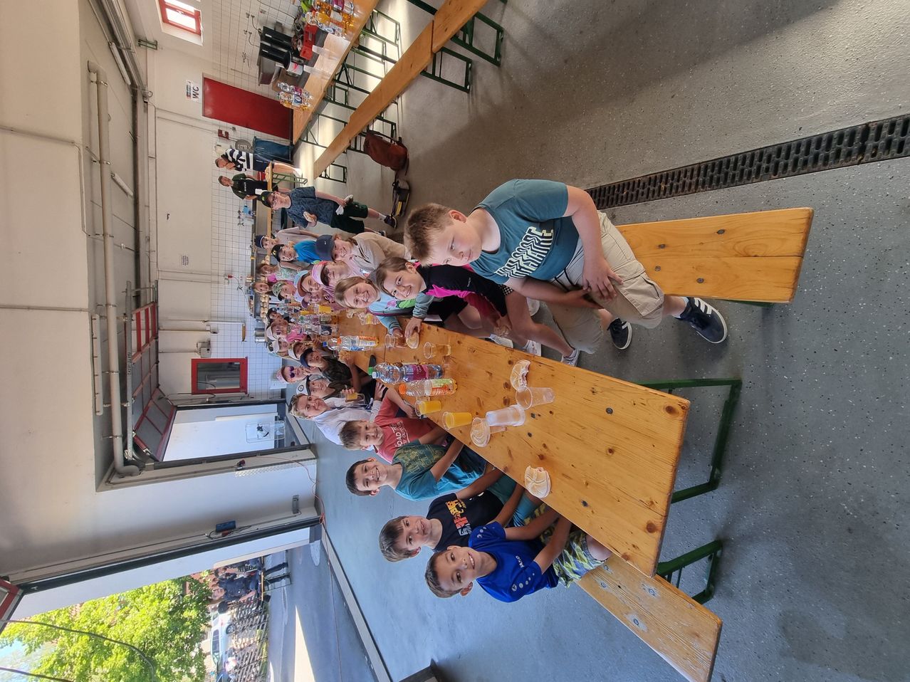 Eine Gruppe von Kindern sitzt mit Getränken um einen Holztisch, lächelt und befindet sich in einem Raum. Es gibt andere Personen im Hintergrund.