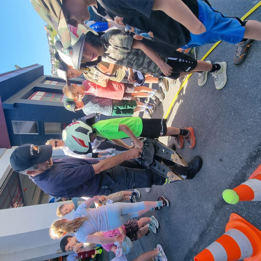 Eine Gruppe von Kindern steht in einer Reihe auf der Straße, einige tragen Helme. Ein Mann in einem blauen Shirt hilft einem Kind in Grün. Verkehrskegel und ein Gebäude sind im Hintergrund.