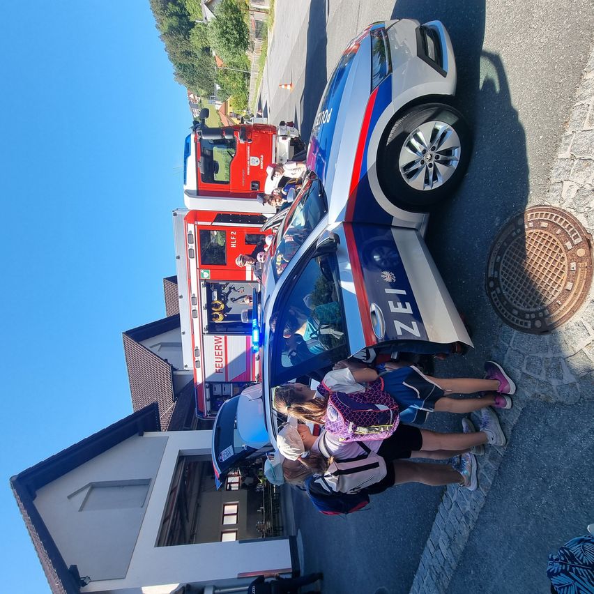 Eine Gruppe von Menschen um ein Polizeiauto und einen Feuerwehrwagen mit der Aufschrift 'Zei' auf dem Polizeiauto. Der Feuerwehrwagen hat das Wort 'Hif' darauf.