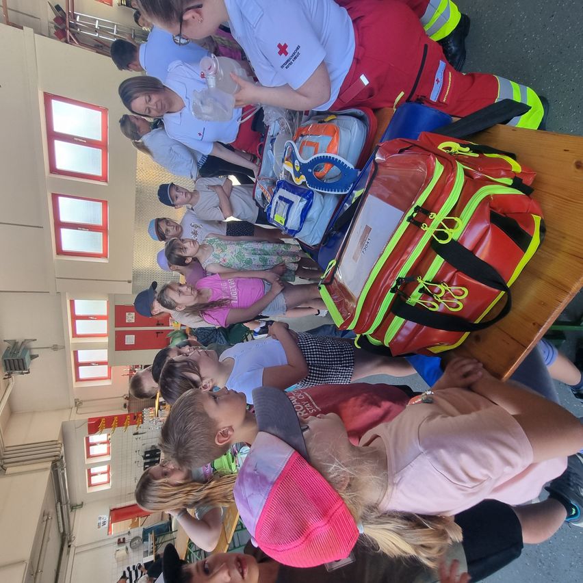 Eine Gruppe von Kindern ist um eine Erste-Hilfe-Station versammelt. Eine erwachsene Person in roter und weißer Uniform kümmert sich um sie. Erste-Hilfe-Ausrüstung steht auf einem Tisch.