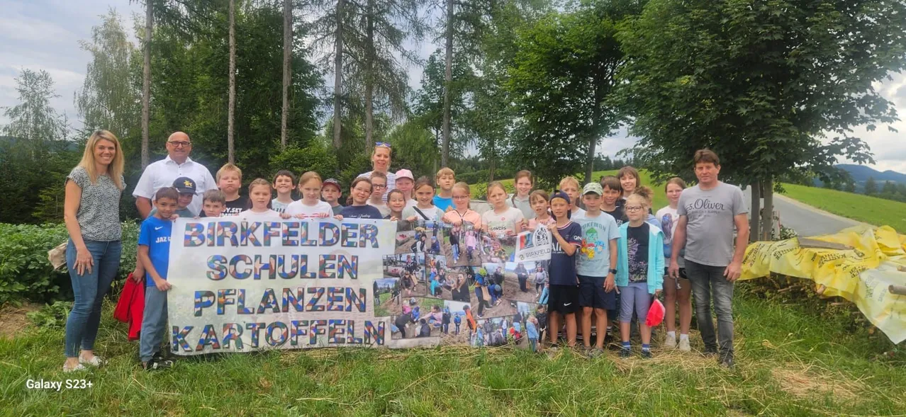 Eine Gruppe von Kindern und Erwachsenen steht hinter einem Banner in einem Feld mit Fotos von Menschen, die Blumen pflanzen. Bäume und Gras sind im Hintergrund.