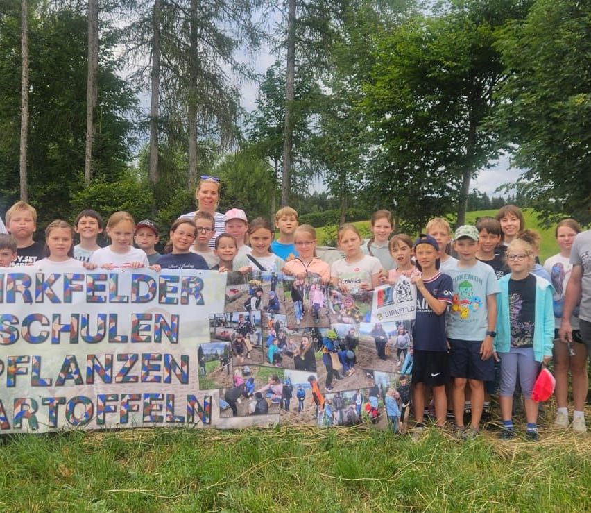 Eine Gruppe von Kindern und Erwachsenen steht hinter einem Banner in einem Feld mit Fotos von Menschen, die Blumen pflanzen. Bäume und Gras sind im Hintergrund.