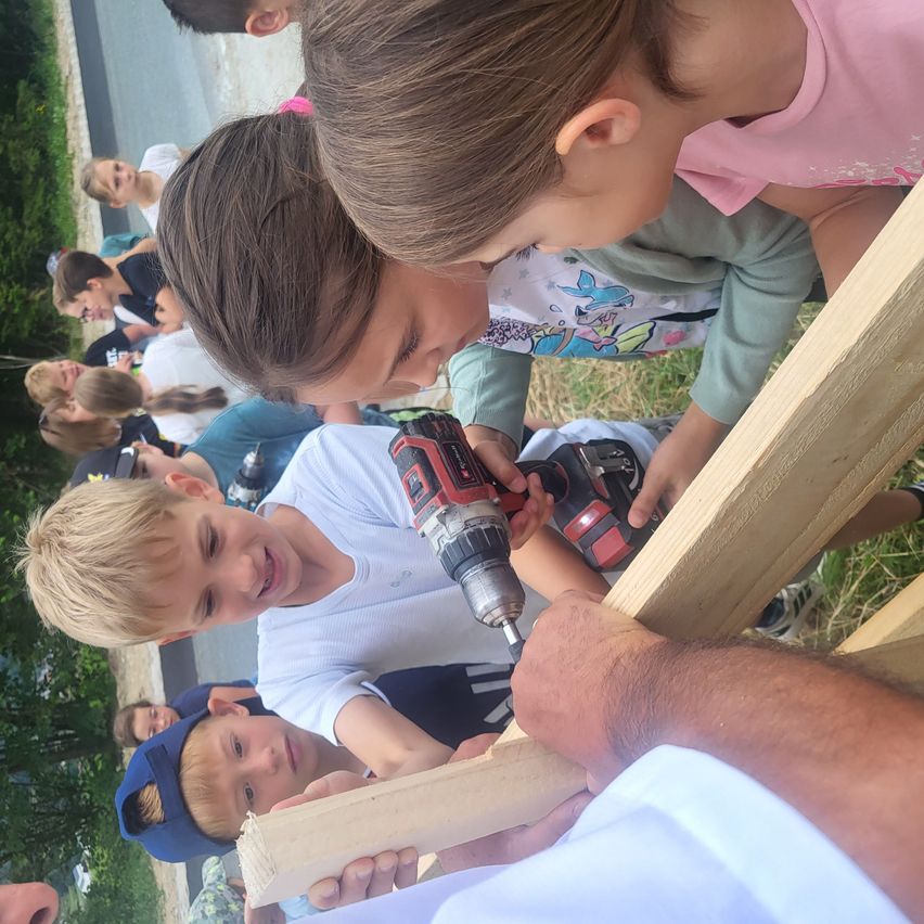 Eine Gruppe von Kindern und einem Erwachsenen bohrt mit einem Bohrer an einem Holzrahmen an einem sonnigen Tag.