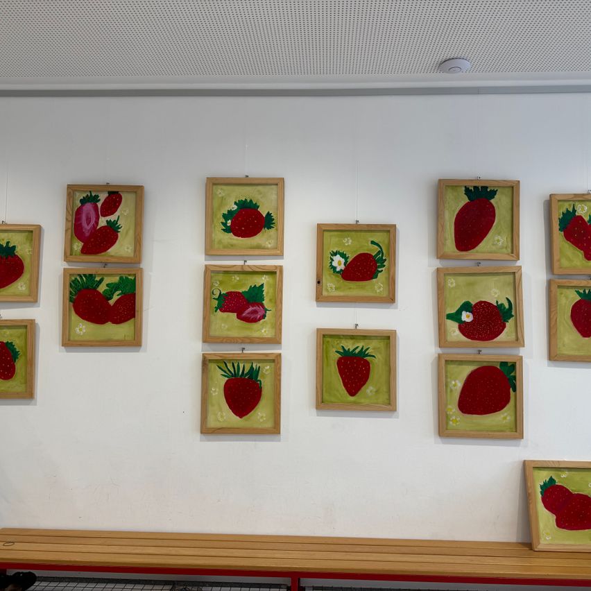 An einer weißen Wand hängen zwölf gerahmte Gemälde von Erdbeeren, wobei ein Gemälde eine Erdbeere mit einer Blume zeigt. Unten steht eine Holzbank.