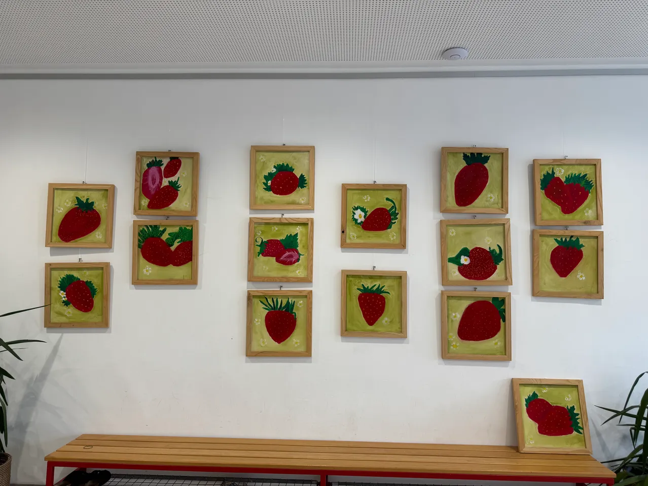 An einer weißen Wand hängen zwölf gerahmte Gemälde von Erdbeeren, wobei ein Gemälde eine Erdbeere mit einer Blume zeigt. Unten steht eine Holzbank.