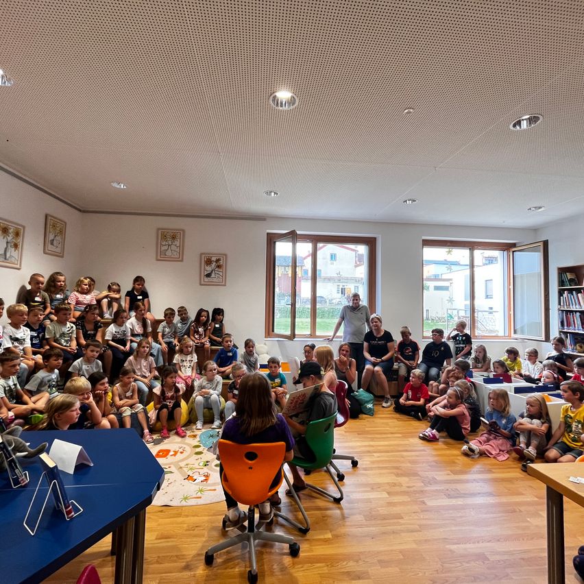 Eine Gruppe von Kindern und Erwachsenen ist in einem Raum versammelt, möglicherweise für eine Lesung. Einige sitzen auf Stühlen, andere stehen. Ein Mann steht in der Nähe des Fensters.