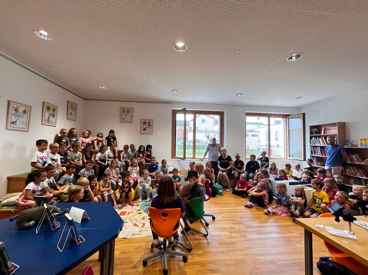 A group of children and adults are gathered in a room, possibly for a reading session. Some are sitting on chairs while others are standing. A man is standing near the window.
