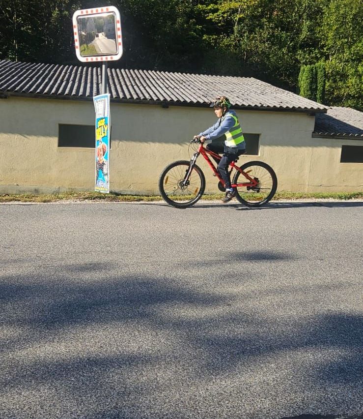 Eine Person fährt auf einem Fahrrad auf einer Straße mit einem Gebäude und Bäumen im Hintergrund.