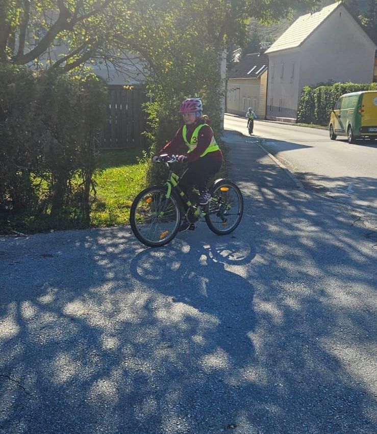 Eine Person fährt auf einem Kiesweg mit einem Fahrrad, mit Bäumen und einem Haus im Hintergrund.