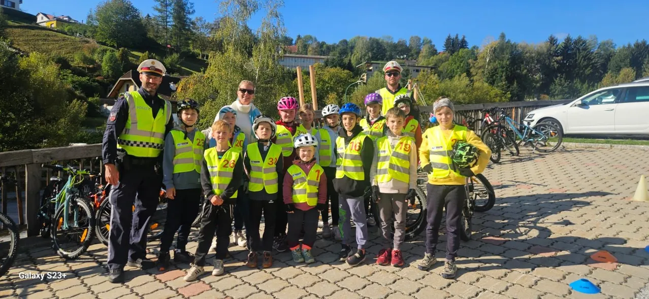 Eine Gruppe von Kindern und Erwachsenen in reflektierenden Westen und Helmen steht auf einem gepflasterten Weg. Sie posieren für ein Foto mit Fahrrädern und einem weißen Auto im Hintergrund.