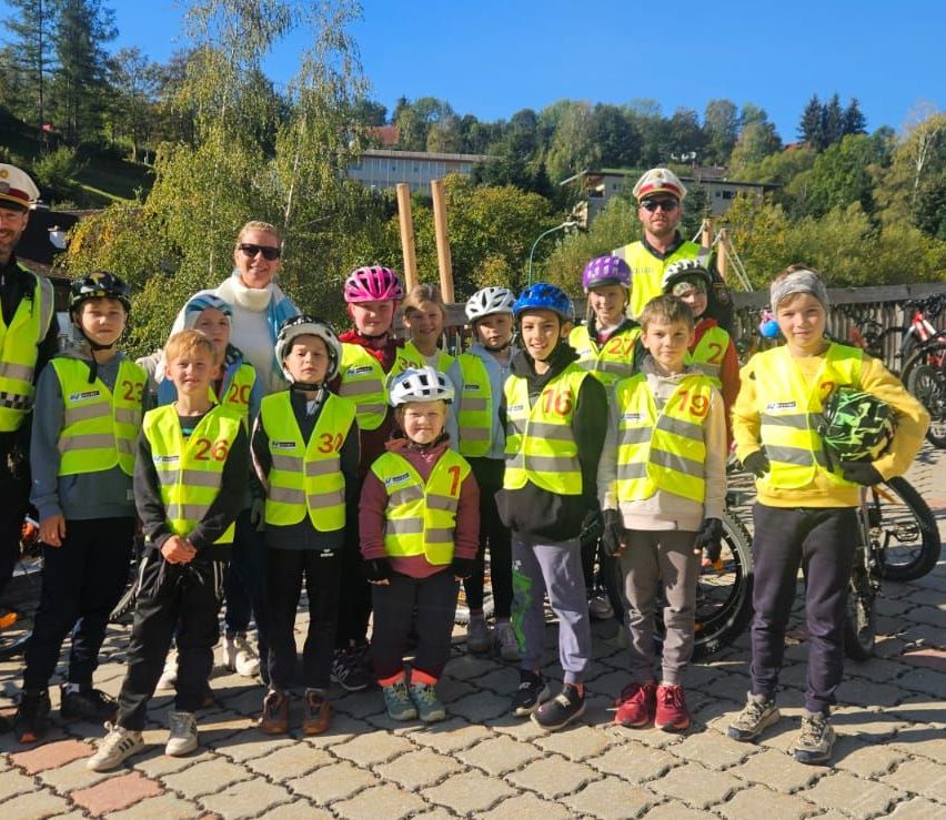 Eine Gruppe von Kindern und Erwachsenen in reflektierenden Westen und Helmen steht auf einem gepflasterten Weg. Sie posieren für ein Foto mit Fahrrädern und einem weißen Auto im Hintergrund.