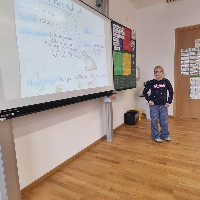 Ein junges Mädchen steht vor einer Tafel in einem Klassenzimmer. Die Tafel hat Zeichnungen und darauf geschriebenen Text.