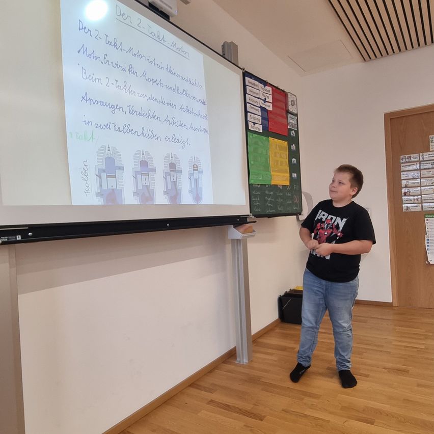 Ein junger Junge steht vor einer Projektionsleinwand in einem Klassenzimmer. Die Leinwand zeigt ein Diagramm einer Maschine mit Text in deutscher Sprache.