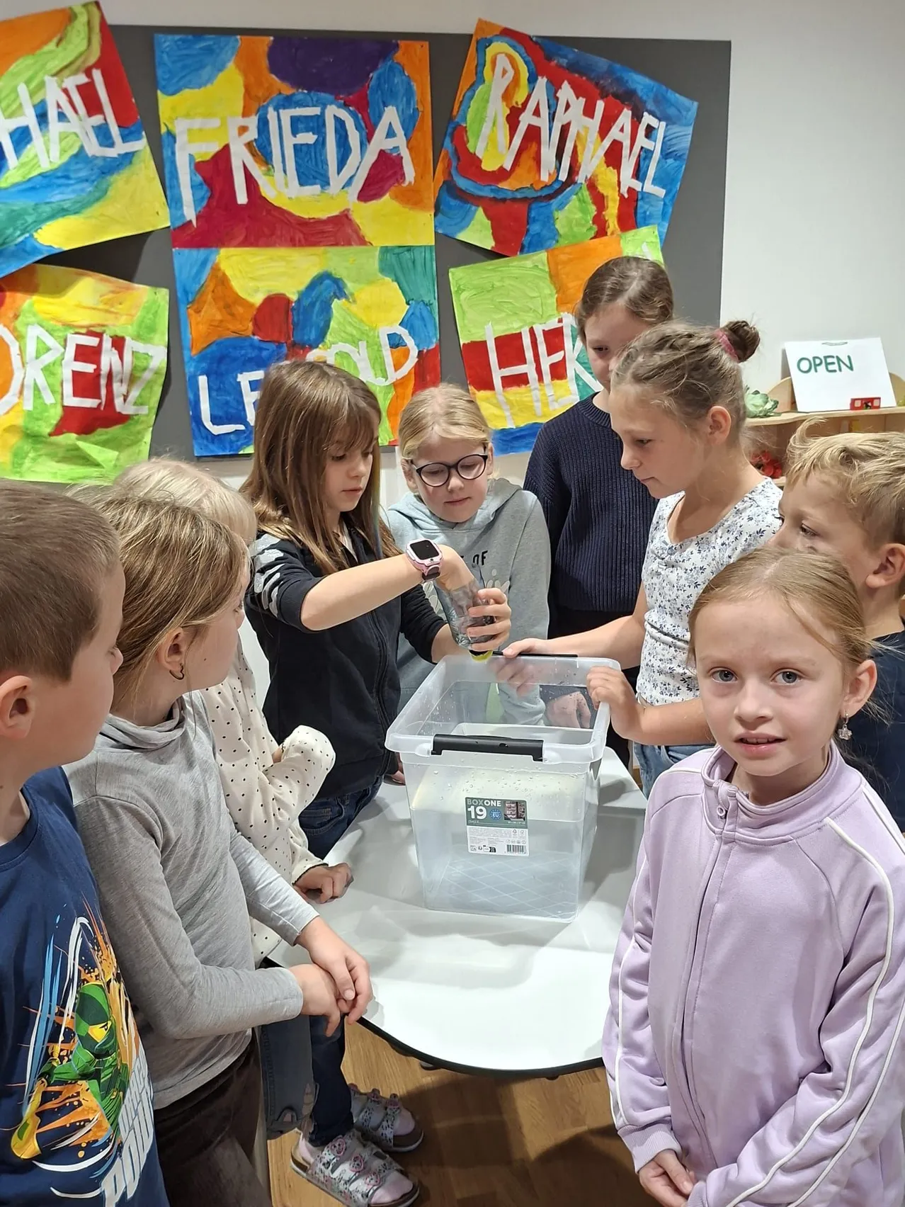 Eine Gruppe von Kindern ist um einen Tisch mit einem Plastikbehälter versammelt. Sie schauen sich etwas im Inneren des Behälters an.