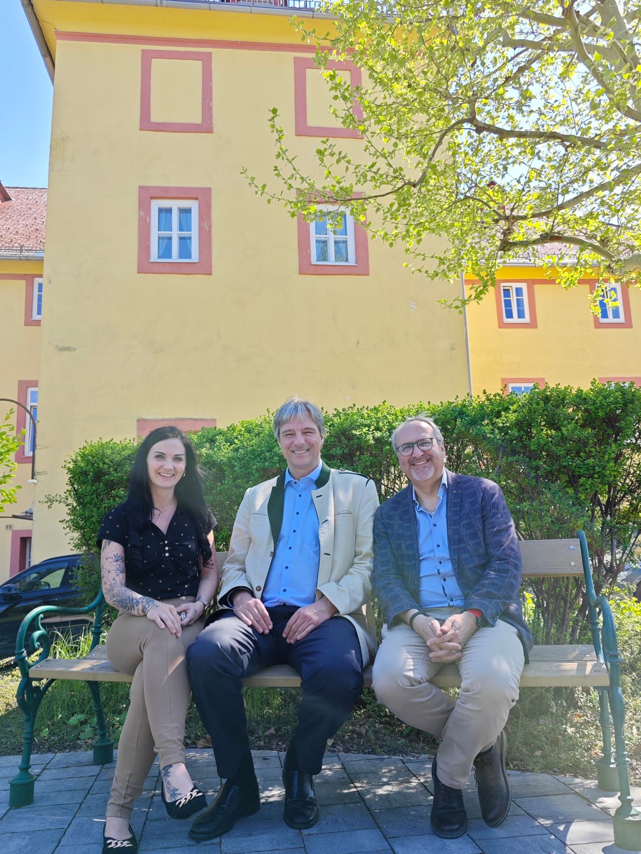 Drei Personen sitzen auf einer Bank vor einem gelben Gebäude. Eine Frau und zwei Männer lächeln und schauen in die Kamera.