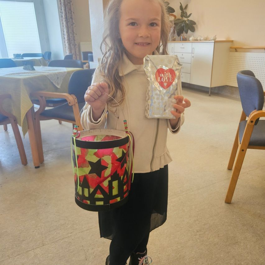 Ein kleines Mädchen steht in einem Raum und hält einen bunten Eimer und eine silberne Tasche mit den Worten 'mit Liebe' darauf. Sie trägt eine weiße Jacke und schwarze Hose.