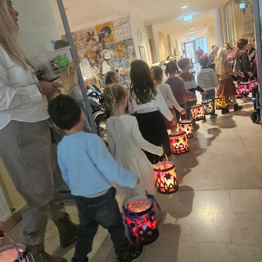 Eine Gruppe von Kindern mit dekorierten Laternen, die in einem Flur gehen, begleitet von Erwachsenen. Eine Frau mit einem Korb steht in der Nähe.
