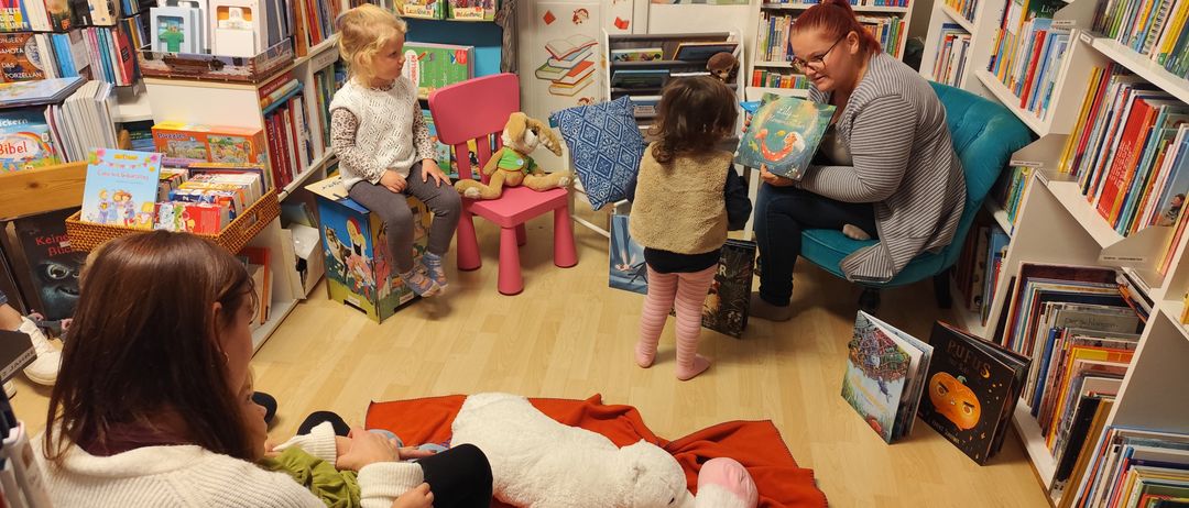 In einer Leseecke sitzen zwei Frauen mit Kindern, eine liest einer kleinen Mädchen in pinken Leggings vor. In der Nähe sitzt ein weiteres Kind auf einem Stuhl und hält ein Kuscheltier. Der Raum ist mit Büchern an den Regalen gefüllt.