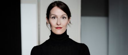 Eine Frau mit kurzen Haaren in einem schwarzen Rollkragenpullover steht mit verschränkten Armen in einem Studio.