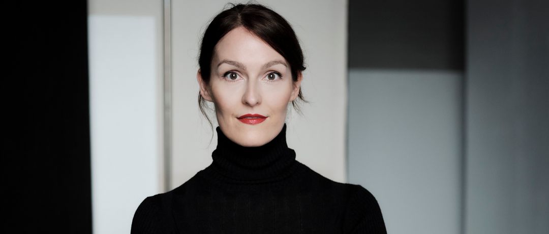 Eine Frau mit kurzen Haaren in einem schwarzen Rollkragenpullover steht mit verschränkten Armen in einem Studio.