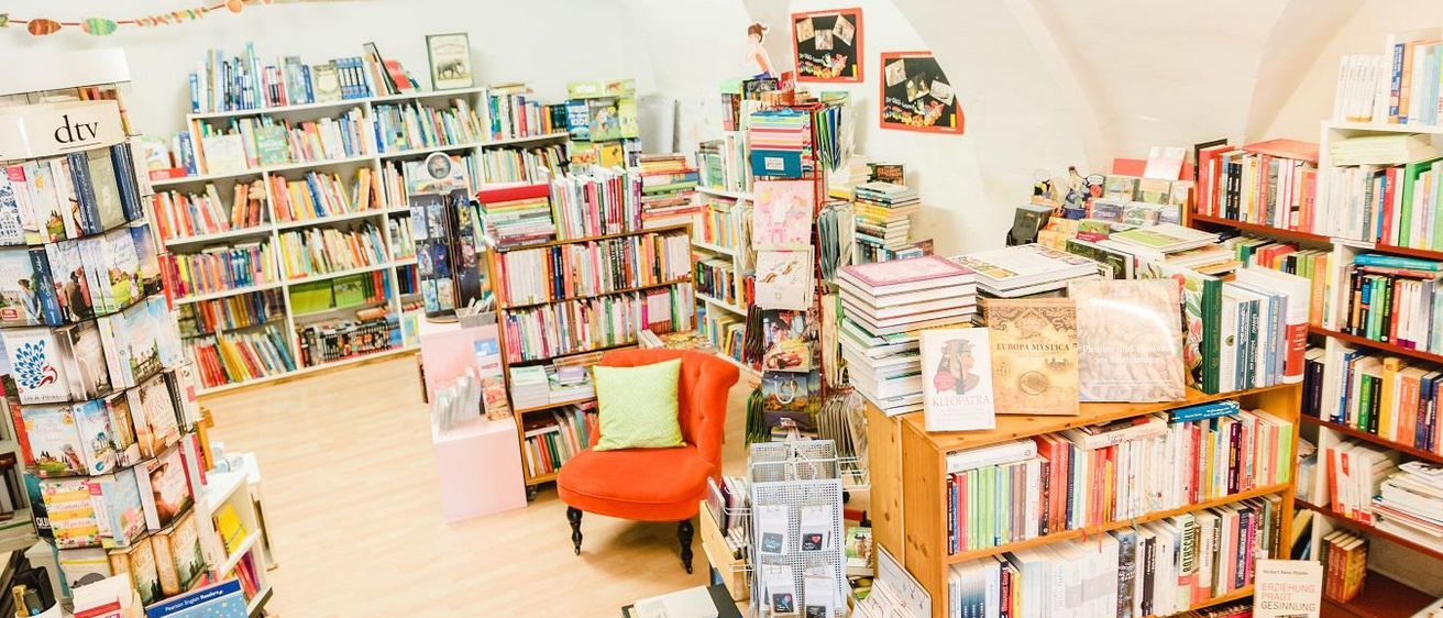 Eine gemütliche Buchhandlung voller Bücherregalen. Ein roter Stuhl mit einem grünen Kissen steht in der Mitte. Die Decke ist gewölbt und ein helles Licht erhellt den Raum.