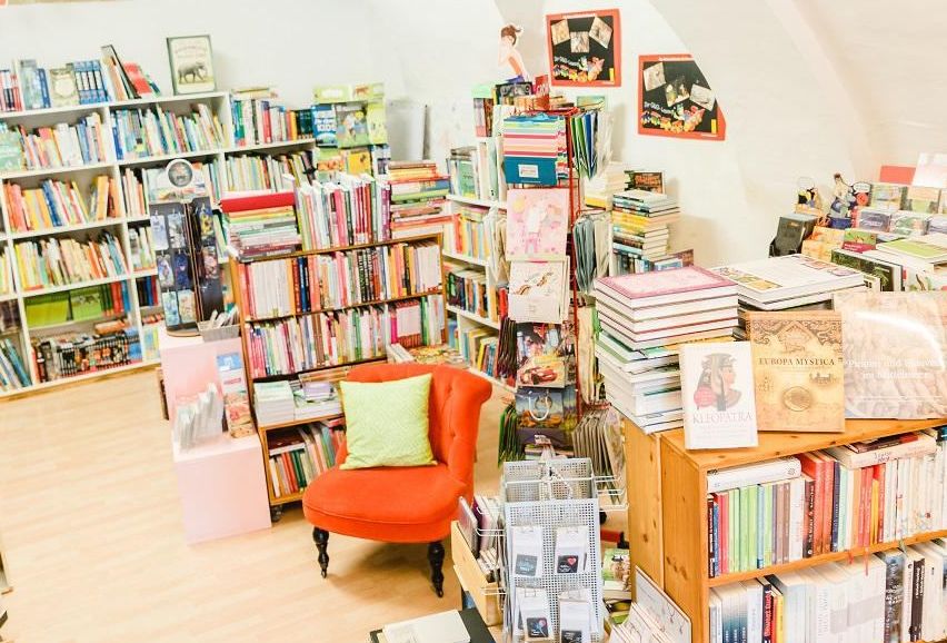 Eine gemütliche Buchhandlung voller Bücherregalen. Ein roter Stuhl mit einem grünen Kissen steht in der Mitte. Die Decke ist gewölbt und ein helles Licht erhellt den Raum.
