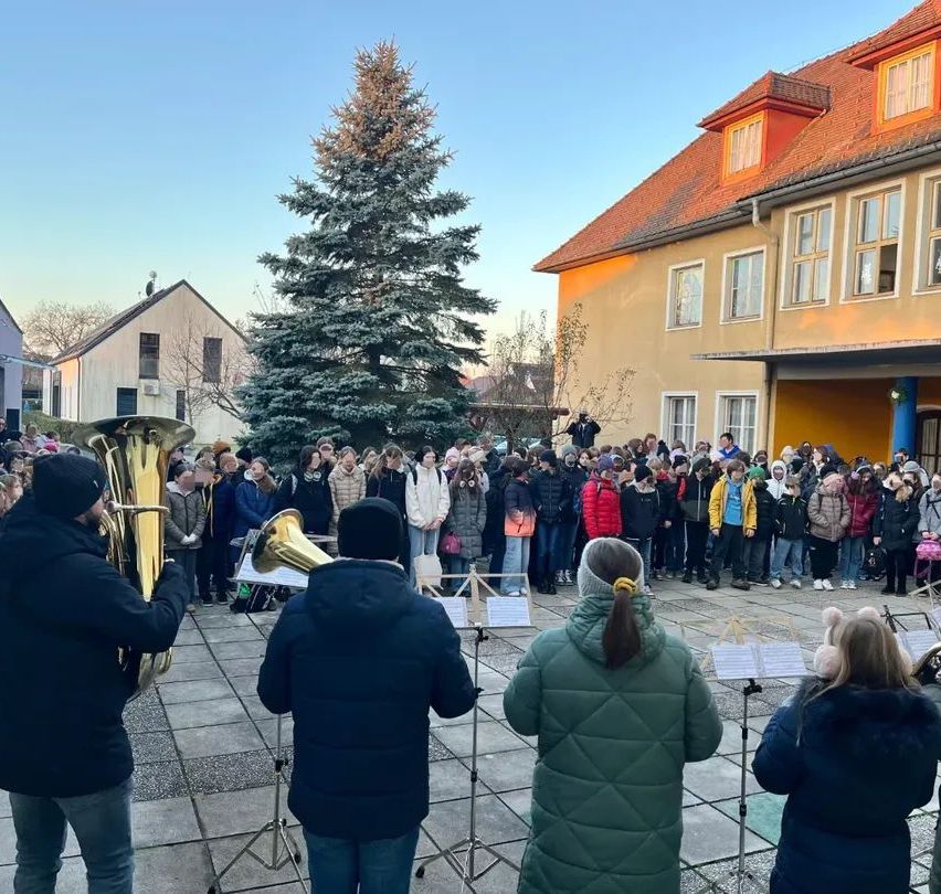 Eine Gruppe von Musikern spielt Instrumente vor einer Menschenmenge, die in einem Innenhof vor einem Gebäude steht, an einem sonnigen Tag.