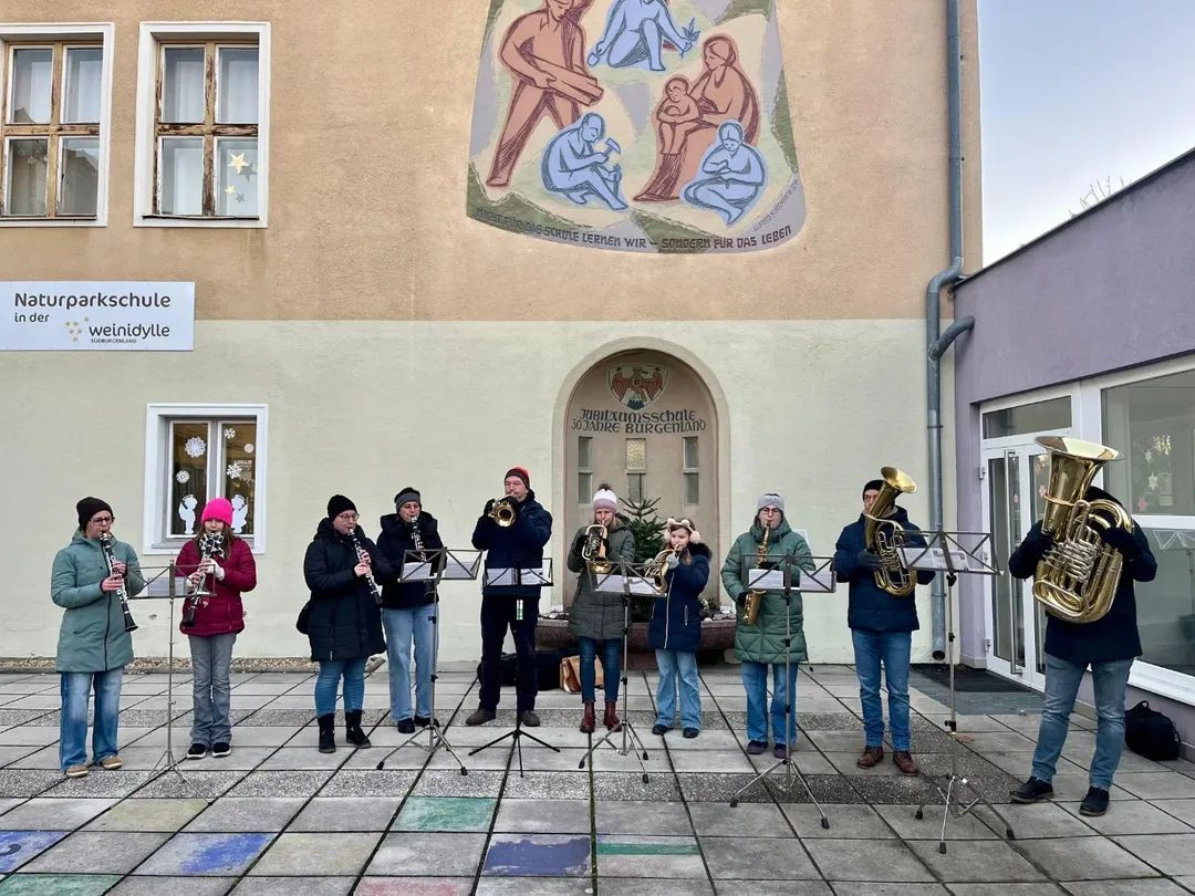 Eine Gruppe von Musikern steht vor einem Gebäude mit einer Wandmalerei. Sie spielen Trompeten, Saxophone und Tuben. Ein Schild am Gebäude lautet 'Wilhelmschule'.