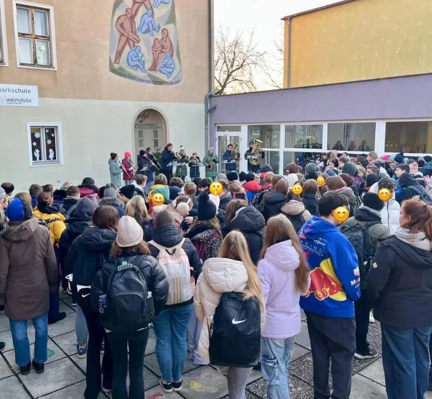 Eine große Menschenmenge, darunter Schüler, versammelt sich vor einem Gebäude. Einige tragen Winterkleidung und haben Rucksäcke dabei. Eine Band tritt in der Nähe des Eingangs auf.