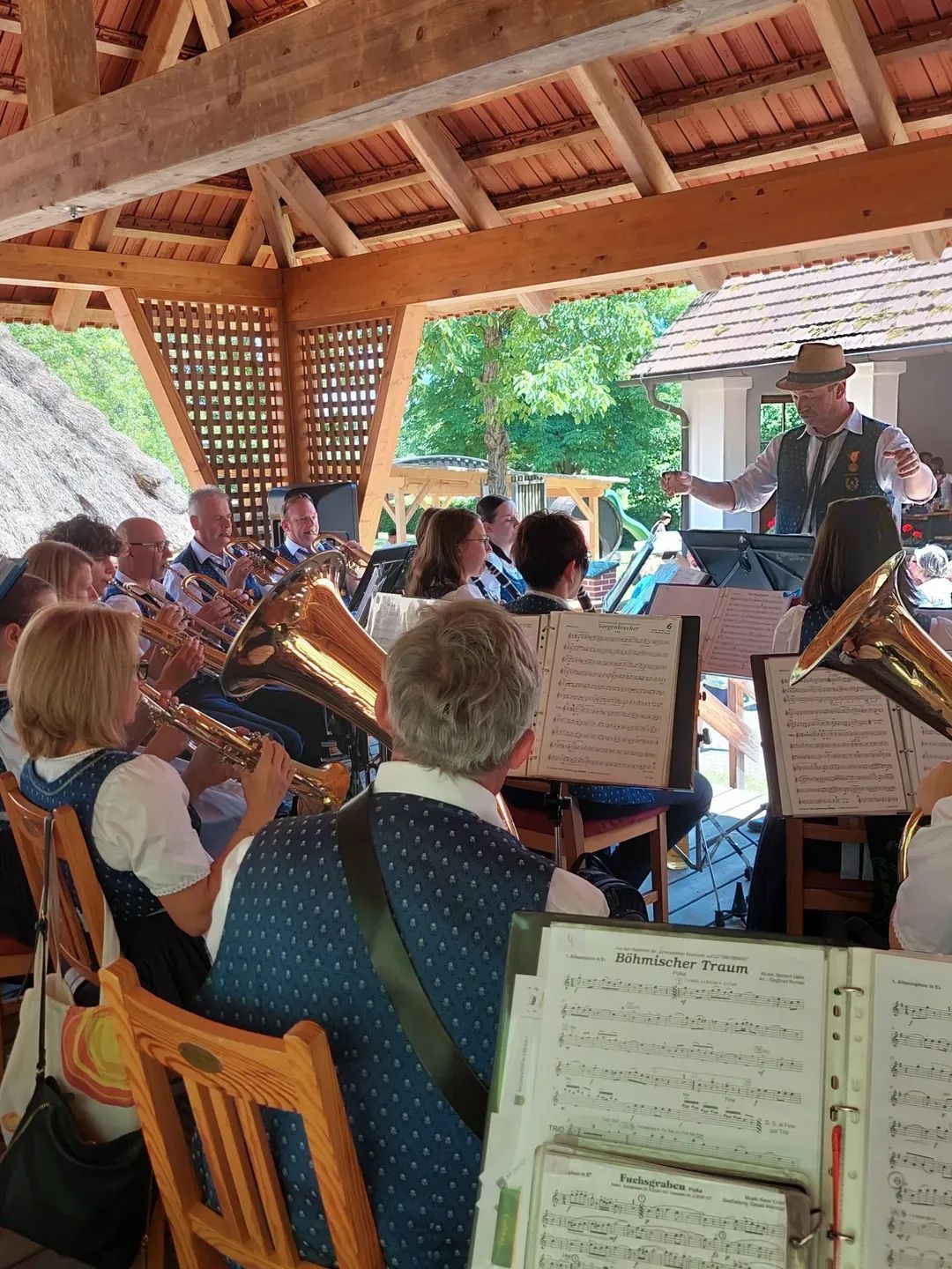 Eine Blechbläsergruppe tritt in einem offenen Pavillon auf. Die Musiker spielen verschiedene Blechblasinstrumente, während ein Dirigent sie leitet. Das Publikum beobachtet die Aufführung von Stühlen aus, die um die Band herum angeordnet sind.