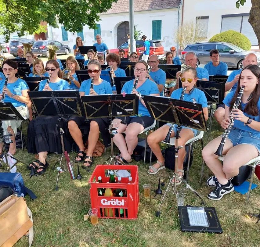 Eine Gruppe von Menschen in blauen T-Shirts spielt Musikinstrumente auf einem Grasfeld. Sie sitzen auf Stühlen mit Notenständern vor sich. Es gibt einen roten Kasten mit Flaschen und einem Becher auf dem Boden.
