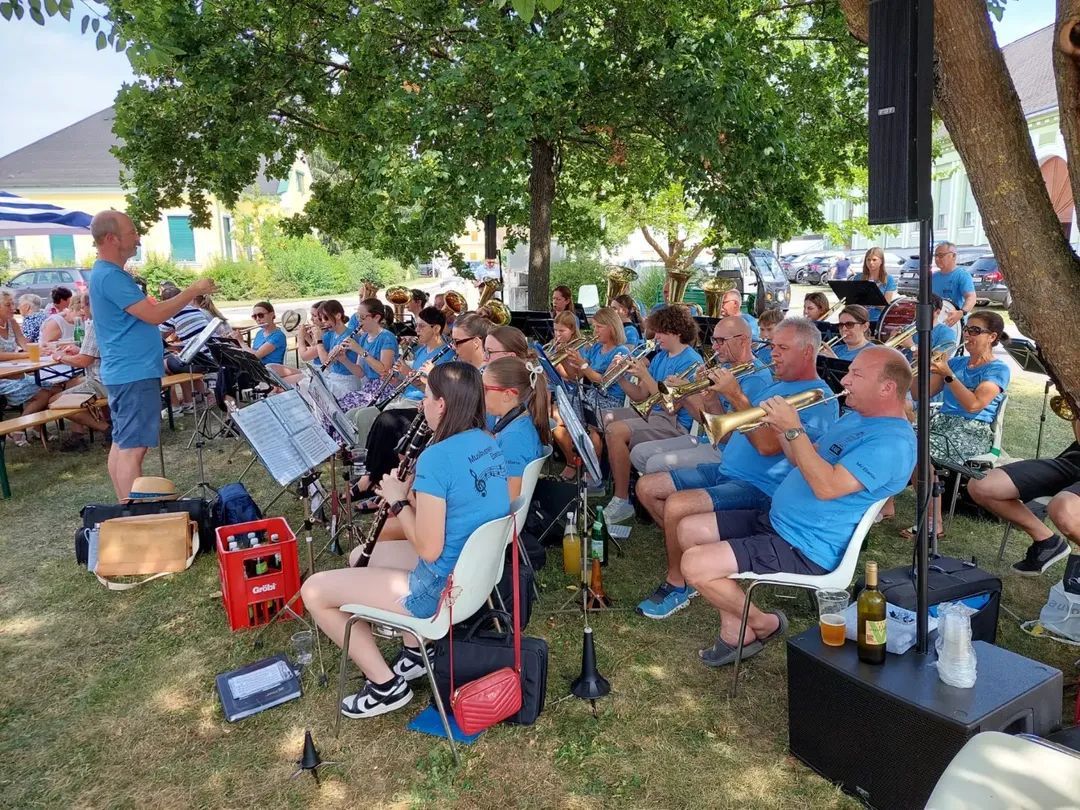 Ein Blasorchester spielt auf einem Rasenfeld unter einem Baum, mit Musikern, die Trompeten, Posaunen und Klarinetten spielen. Einige sitzen auf Stühlen, andere stehen. Musikinstrumente und Noten sind verstreut.