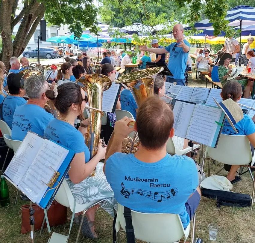 Ein Blechbläserensemble probt im Freien unter einem Baum, während das Publikum auf Stühlen sitzt. Der Dirigent steht vor der Band mit Notenblättern.