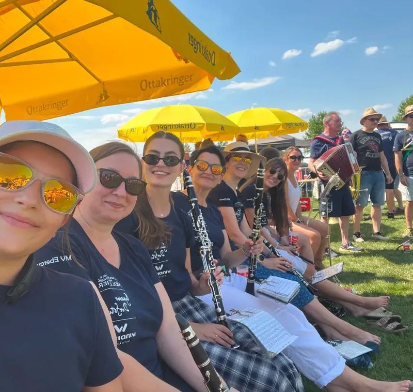 A group of women wearing sunglasses and holding clarinets sit on a grassy field under yellow umbrellas. In the background, there are people standing and sitting, and one person is playing an accordion.