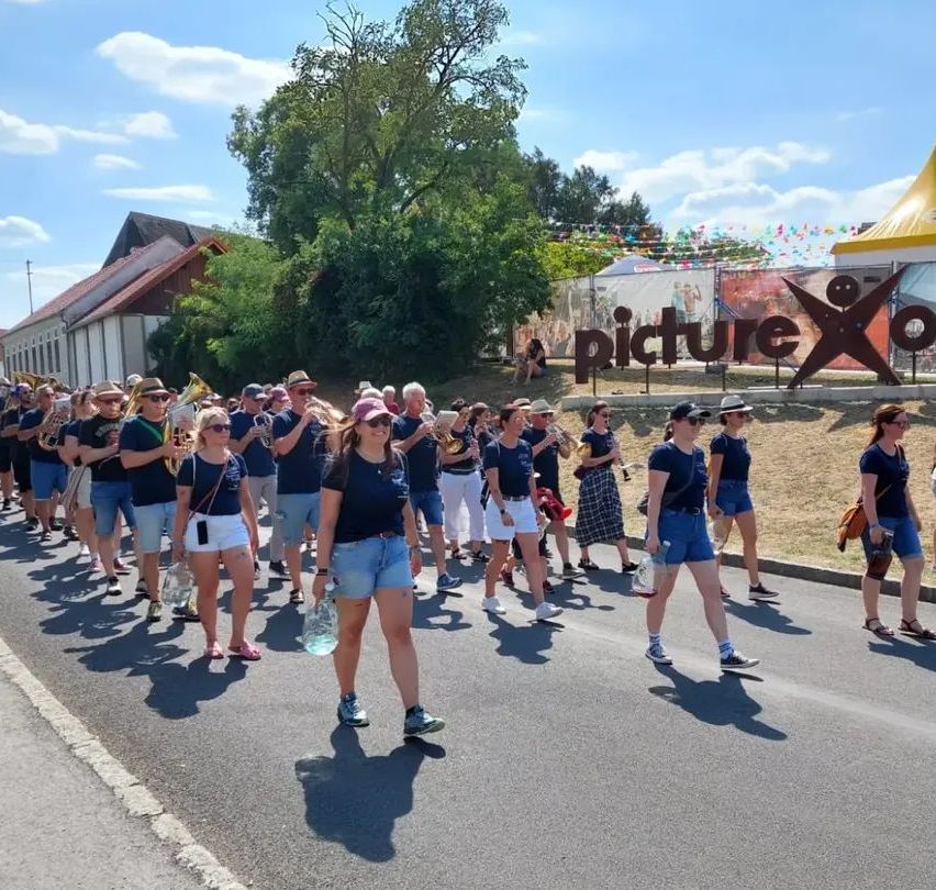 A group of people in matching blue shirts and shorts are marching in a parade, playing musical instruments. They are walking down a road with a large sign that reads 'picture X'.