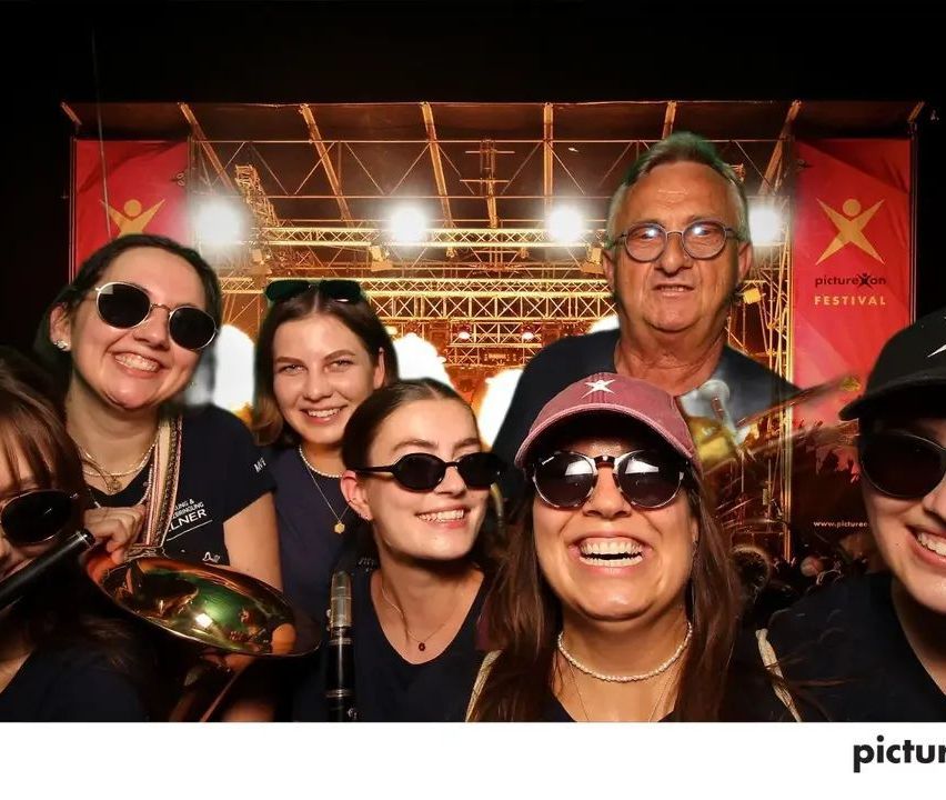 A group of people, including women and a man, are smiling and posing for a photo at a festival with a stage and red banners in the background.