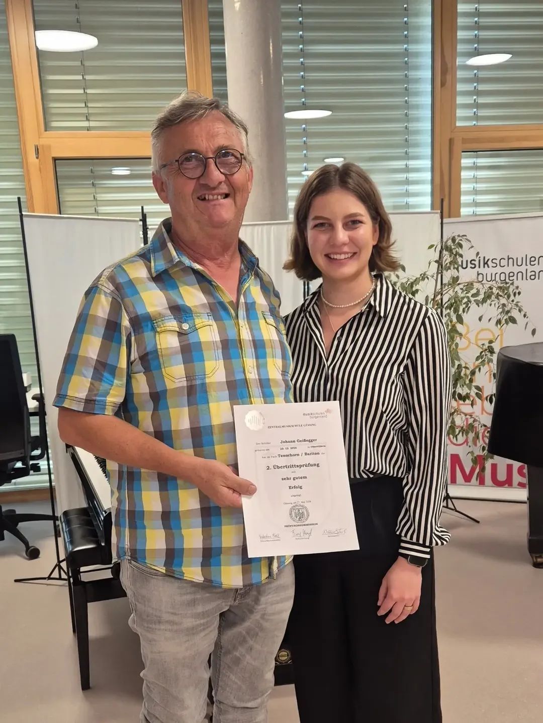 Ein Mann und eine Frau lächeln und halten ein Zertifikat. Der Mann trägt eine Brille und ein kariertes Hemd. Die Frau trägt eine Halskette und ein schwarz-weiß gestreiftes Hemd. Dahinter steht ein Klavier und eine Pflanze. Es gibt auch Banner mit Texten und Logos.