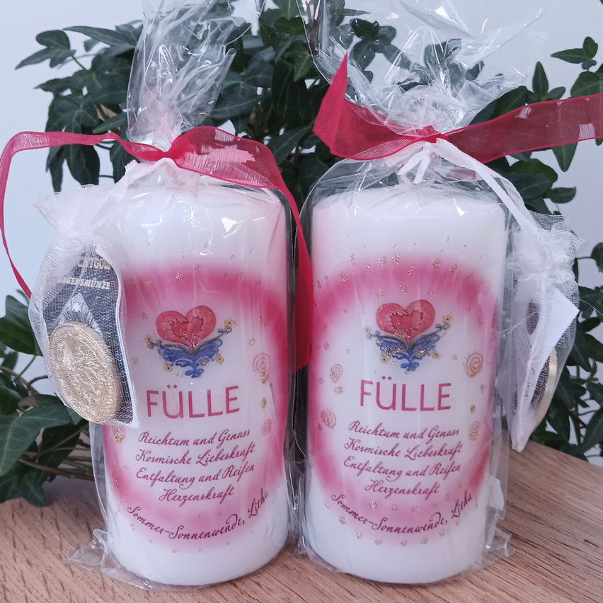 Zwei Kerzen in Plastikverpackung mit einem roten Band, verziert mit pinken und weißen Designs und dem Wort 'Fülle'. Eine Münze ist an der linken Kerze befestigt. Dahinter befinden sich grüne Blätter.
