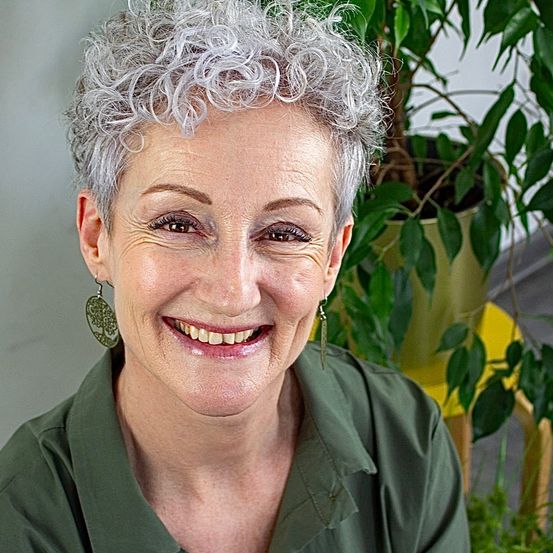 Eine Frau mit grauem Haar lächelt vor einer grünen Pflanze.