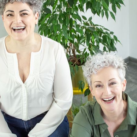 Zwei Frauen lächeln in einem Raum mit einer Pflanze hinter ihnen. Die Frau auf der linken Seite trägt ein weißes Oberteil und die Frau auf der rechten Seite trägt ein grünes Hemd.