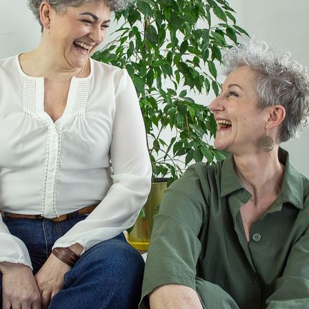 Zwei Frauen sitzen vor einer Pflanze, eine trägt ein weißes Hemd und die andere ein grünes Shirt.