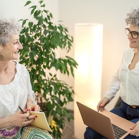 Zwei Frauen sitzen in einem Raum mit einer Pflanze. Eine Frau lächelt und hält ein Notizbuch und einen Stift, die andere schaut auf einen Laptop.
