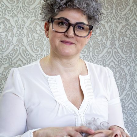 Eine Frau mit grauen Haaren, die eine Brille und eine weiße Bluse trägt und ein kleines graues Objekt in der Hand hält. Die Wand hinter ihr hat ein grau-weißes Blumenmuster.