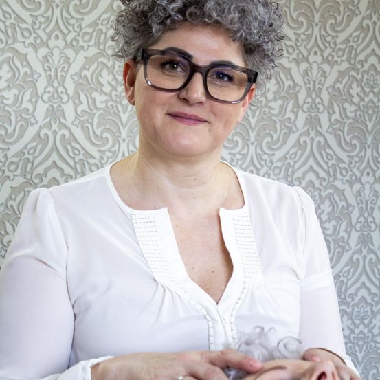 Eine Frau mit grauen Haaren, die eine Brille und eine weiße Bluse trägt und ein kleines graues Objekt in der Hand hält. Die Wand hinter ihr hat ein grau-weißes Blumenmuster.