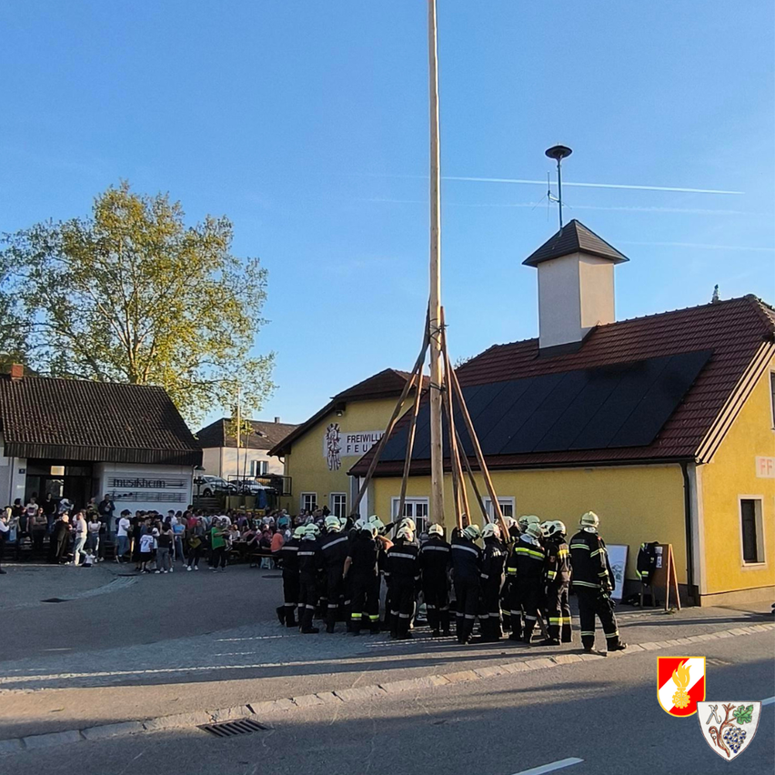 Eine Gruppe von Feuerwehrleuten steht vor einem Gebäude mit vielen Zuschauern. Die Feuerwehrleute halten eine lange Stange in der Mitte der Straße.